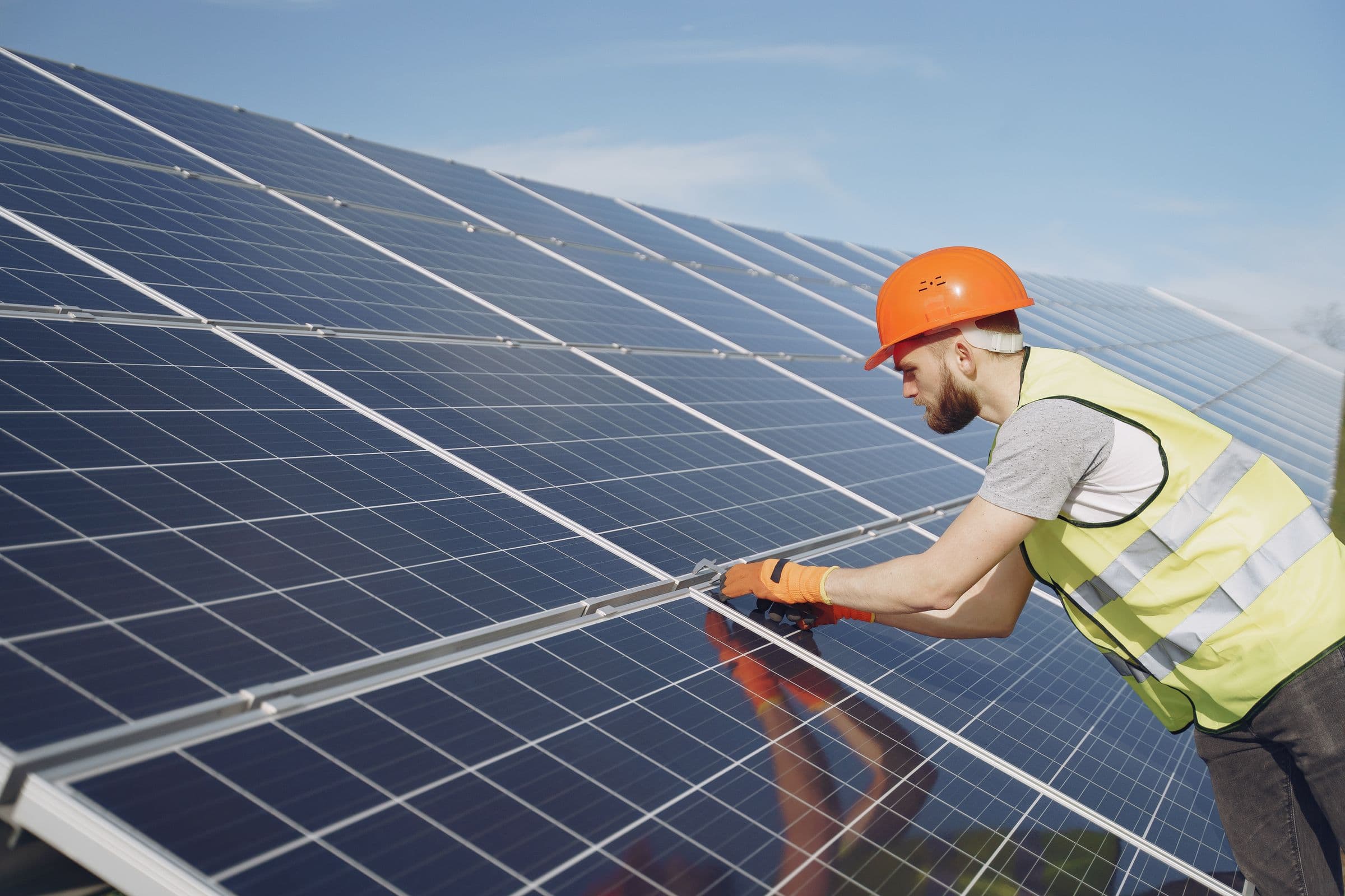 Technicien en gilet fluo ajustant un rail de fixation sur une rangée de panneaux solaires en plein soleil.