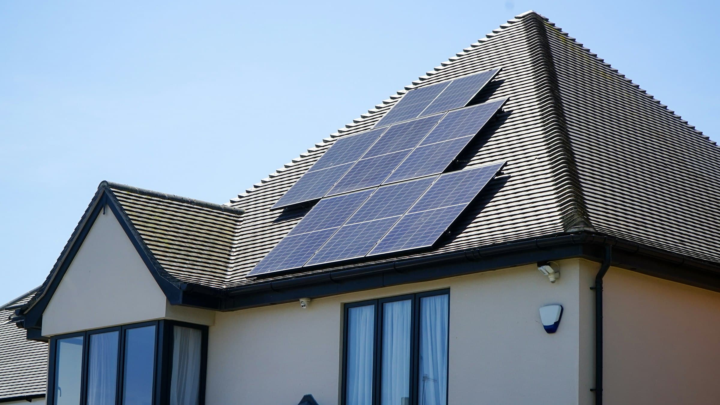 Maison avec toit en tuiles grises et rangée de panneaux solaires noirs, lucarne, ciel légèrement voilé.