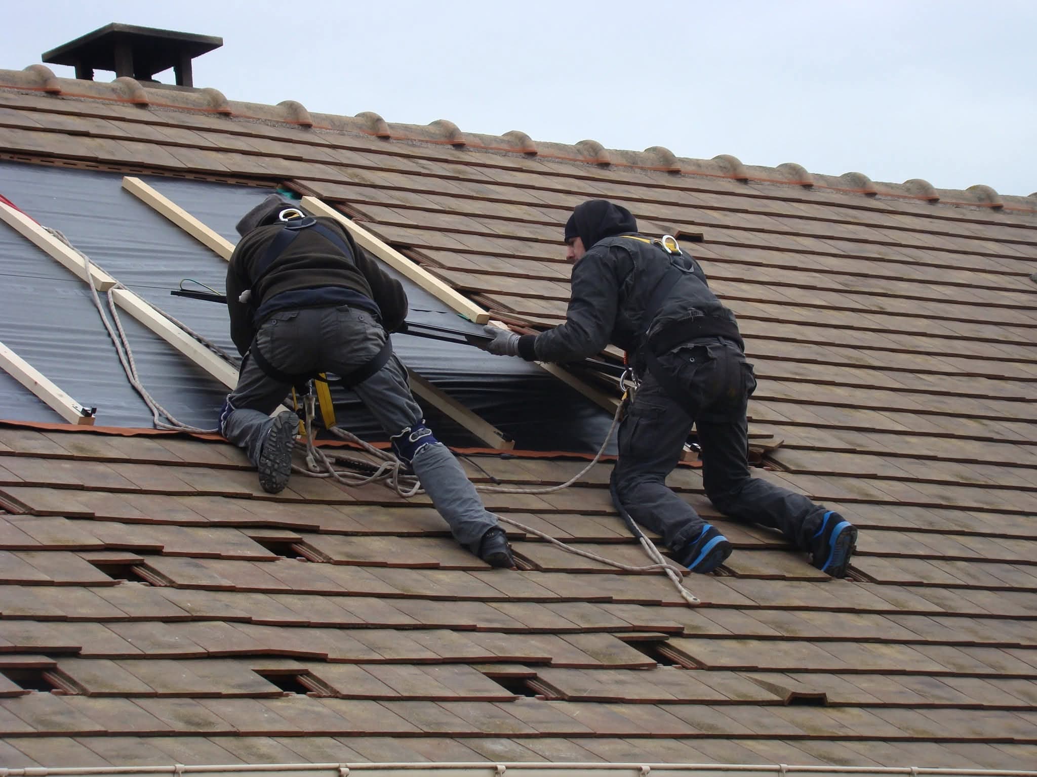 Deux poseurs harnachés et encordés, à genoux sur une toiture en tuiles partiellement découverte, glissant à deux un panneau solaire sous les liteaux.