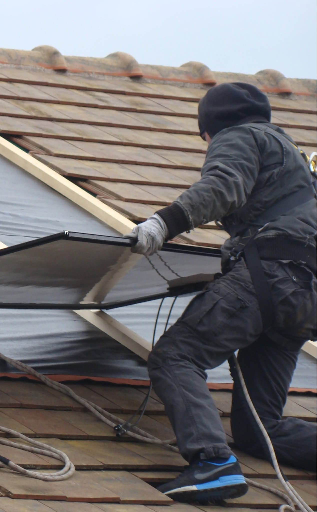 Poseur harnaché, un genou à terre sur une toiture en tuiles dégarnie, engageant un panneau solaire sous les liteaux, corde d’assurage en pied.