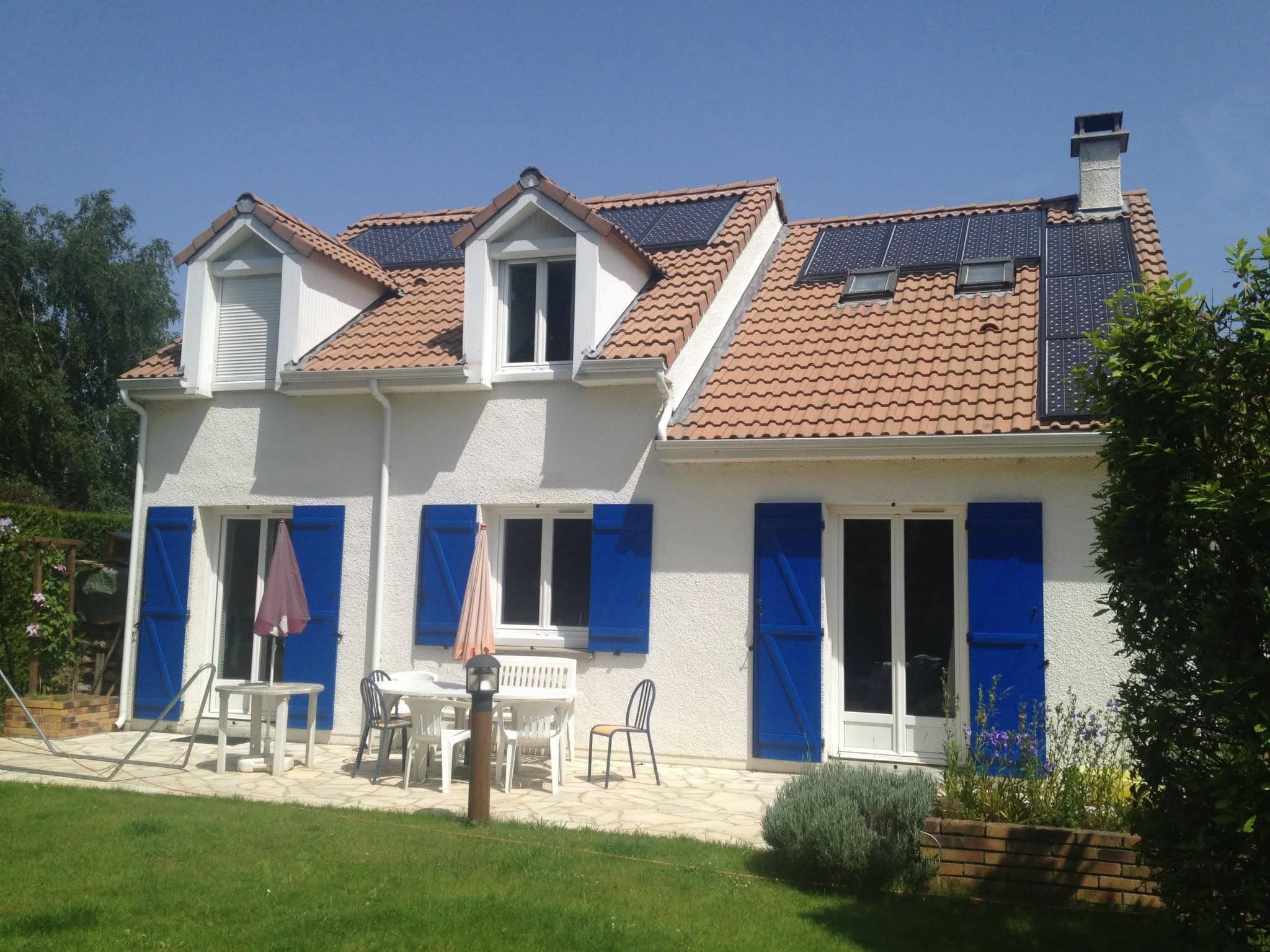 Pavillon crépi blanc à volets bleus, deux lucarnes et cheminée, panneaux solaires posés sur les deux pans de la toiture en tuiles, terrasse et jardin au premier plan.