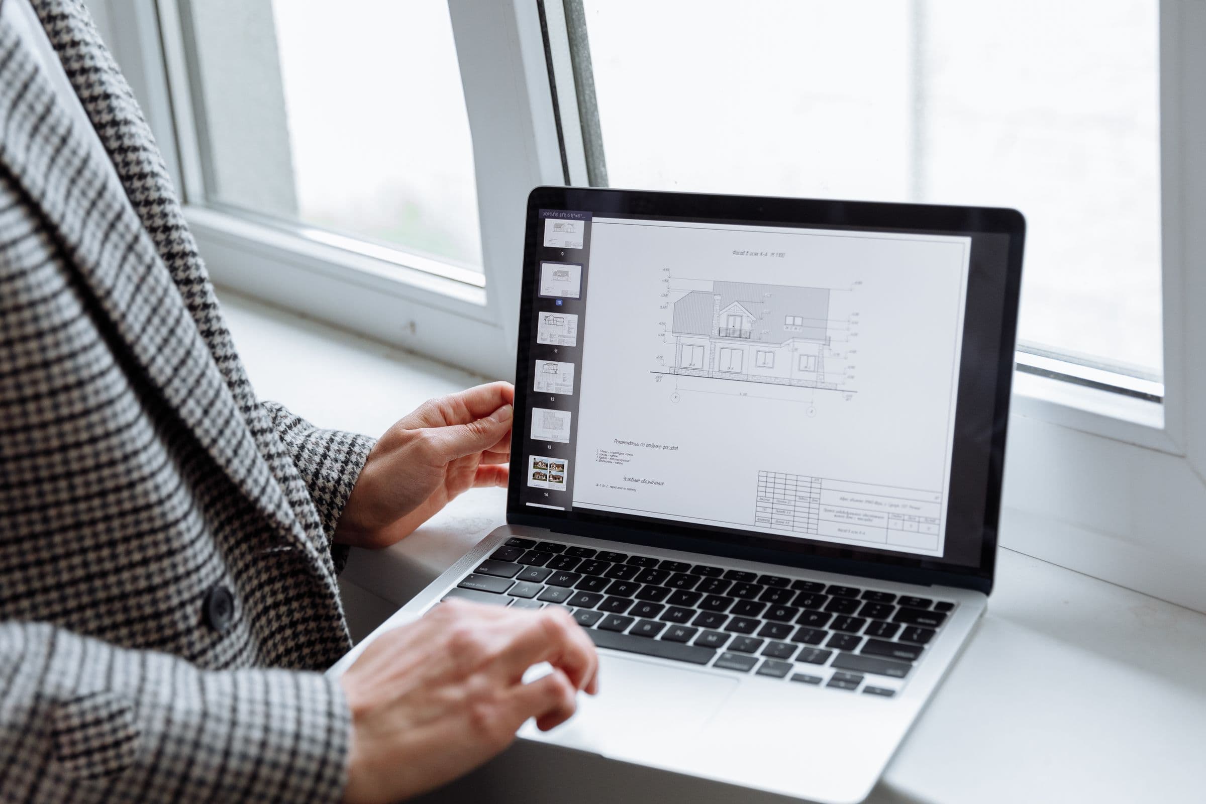Plan coté d’une maison affiché à l’écran d’un ordinateur portable, une main sur le clavier près d’une fenêtre.