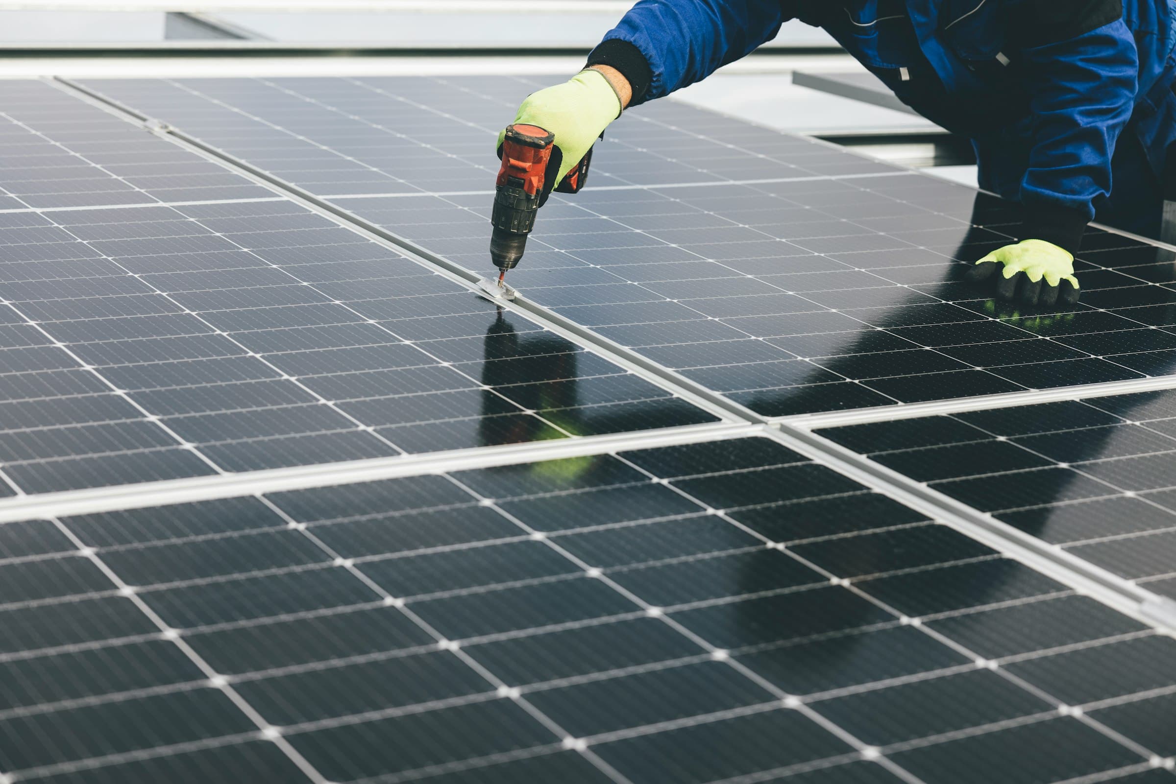 Gros plan d'un technicien qui visse un rail de fixation de panneau solaire avec une visseuse électrique, gants de protection.