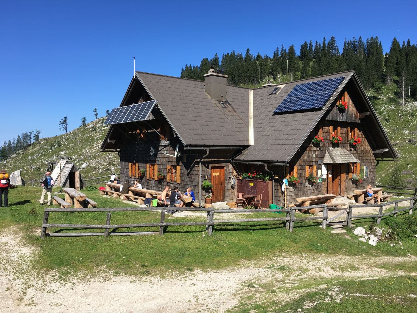 Chalet d’alpage en bardeaux de bois, deux champs de panneaux solaires sur le toit et sur un support incliné, prairie et sapins.