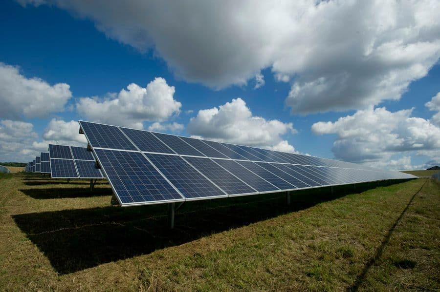 Rangées de panneaux photovoltaïques au sol sous un grand ciel bleu chargé de nuages blancs, vue en contre-plongée.